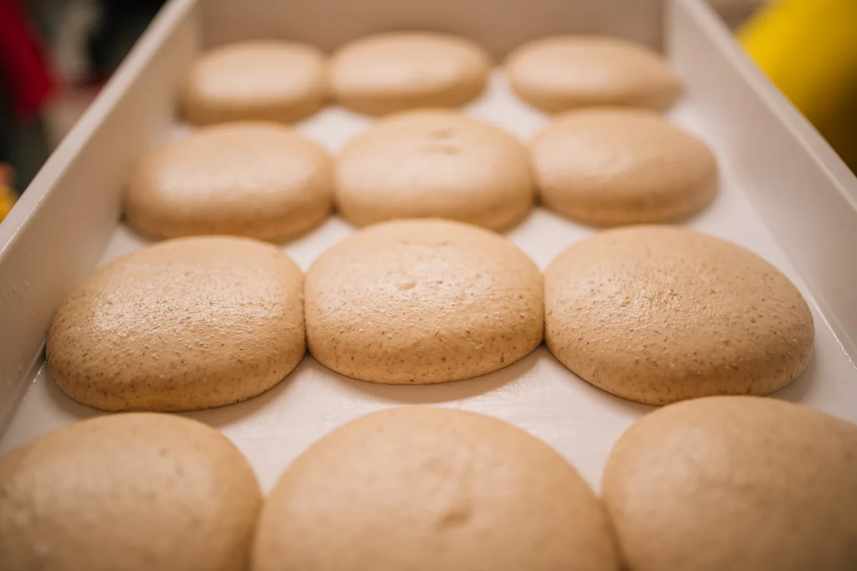 Le mani di un panettiere lavorano l'impasto Polesanissimo® tra panetti lievitati e farina in una luce calda.